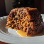 A close-up of a rich, dark slice of plum pudding covered in caramel sauce on a white plate.