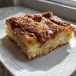 A square slice of baked french toast with a rich, caramelized cinnamon sugar topping on a white plate.