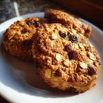 Three freshly baked oatmeal raisin cookies piled on a white plate, showing visible oats and dark raisins.