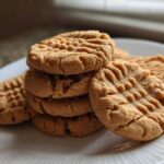 A stack of freshly baked peanut butter cookies featuring the classic crisscross fork pattern, resting on a white plate.