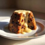A close-up of a moist slice of plum pudding, studded with raisins, topped with a cascade of rich vanilla custard sauce.