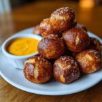 A pile of freshly baked, golden brown pretzel bites sprinkled with coarse salt, served next to a small bowl of bright orange cheese sauce.