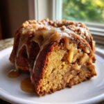 A close-up of a slice of moist apple bundt cake topped with caramel sauce and chunks of apple.