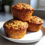 Three freshly baked banana oatmeal muffins stacked on a white plate, featuring a golden-brown, textured top.