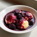 Close-up of warm Blackberry Dumplings served in a white bowl, generously covered in dark purple blackberry sauce and whole berries.