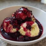 Three fluffy, pale yellow Blackberry Dumplings generously covered in a dark, rich blackberry sauce in a white bowl.