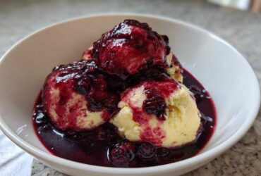 Three fluffy, pale yellow Blackberry Dumplings generously covered in a dark, rich blackberry sauce in a white bowl.