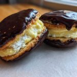 Two sandwich-style boston cream pie cookies filled with vanilla cream and topped with rich chocolate ganache.