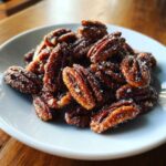 A close-up of freshly made candied pecans recipe nuts piled on a small white plate, glistening with sugar crystals.