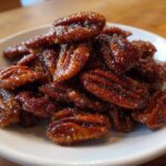 A close-up shot of a pile of glossy, caramelized candied pecans recipe served on a small white plate.