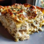 A close-up of a thick slice of chicken alfredo lasagna showing layers of pasta, shredded chicken, and creamy sauce, topped with melted cheese and herbs.
