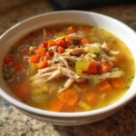 Close-up of a white bowl filled with rich chicken vegetable soup, featuring shredded chicken and diced carrots and celery.
