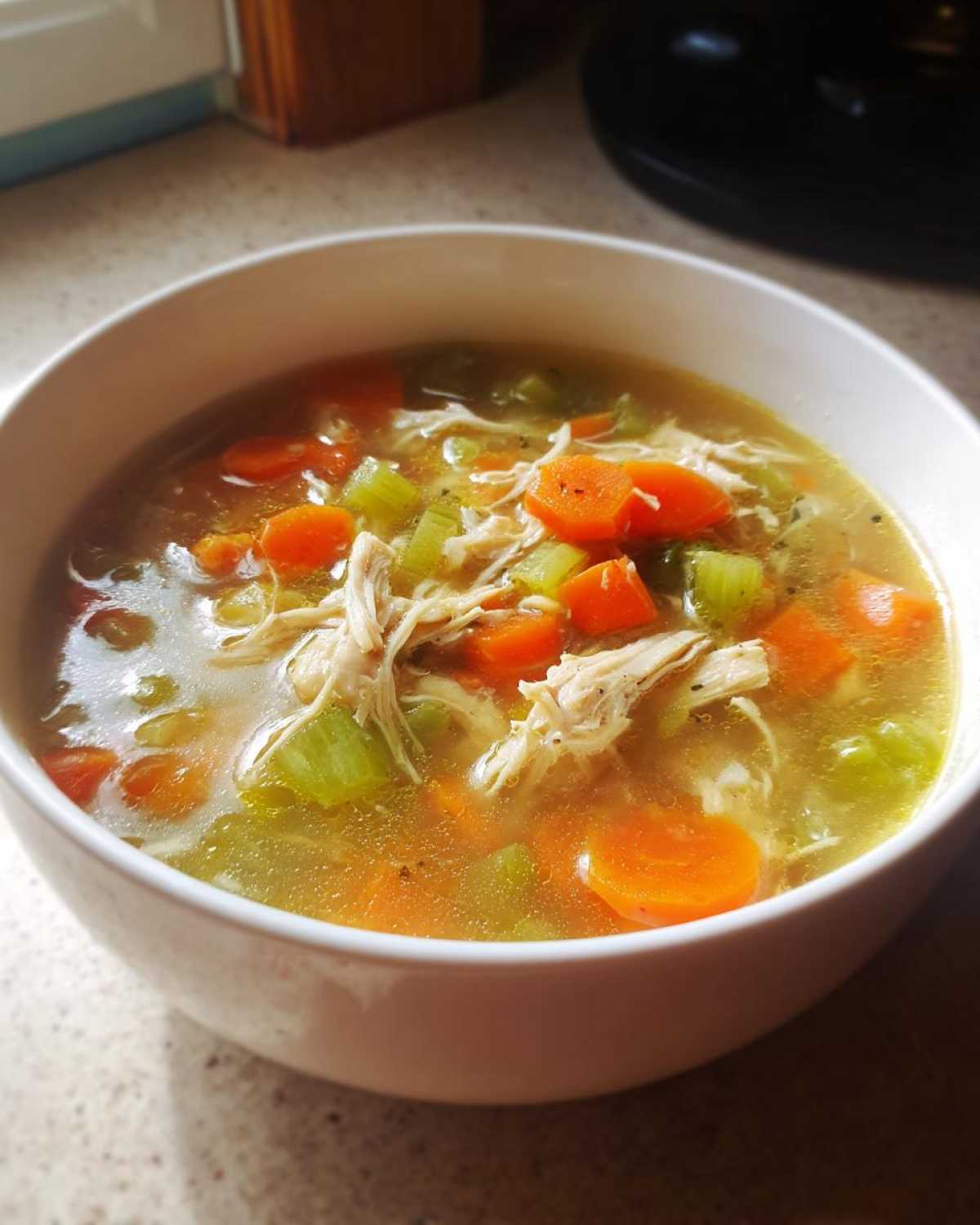 Close-up of a white bowl filled with steaming chicken vegetable soup, featuring shredded chicken, bright orange carrots, and green celery.
