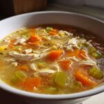 Close-up of a steaming bowl of chicken vegetable soup with shredded chicken, bright orange carrots, and celery chunks.