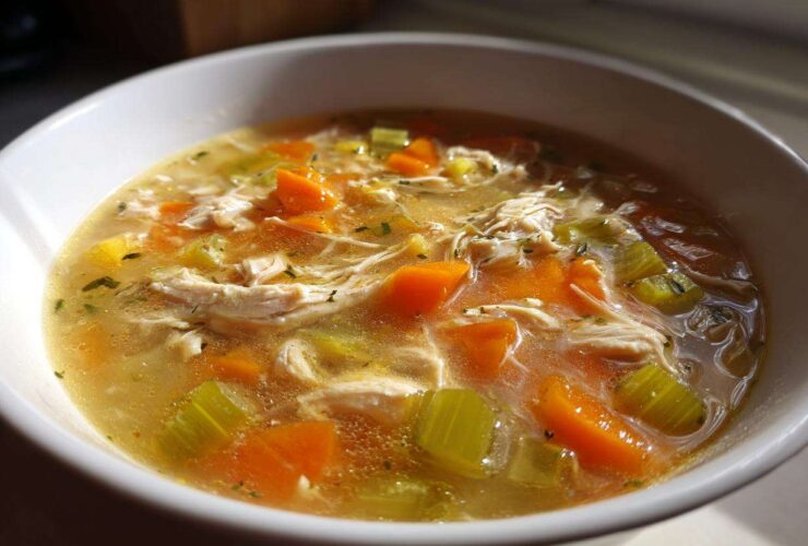 Close-up of a steaming bowl of chicken vegetable soup with shredded chicken, bright orange carrots, and celery chunks.