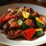 Close-up of glazed chicken zucchini stir fry with red peppers and sesame seeds on a white plate.