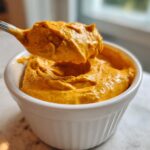 A silver spoon lifting a thick scoop of bright orange beer cheese dip from a white ramekin.