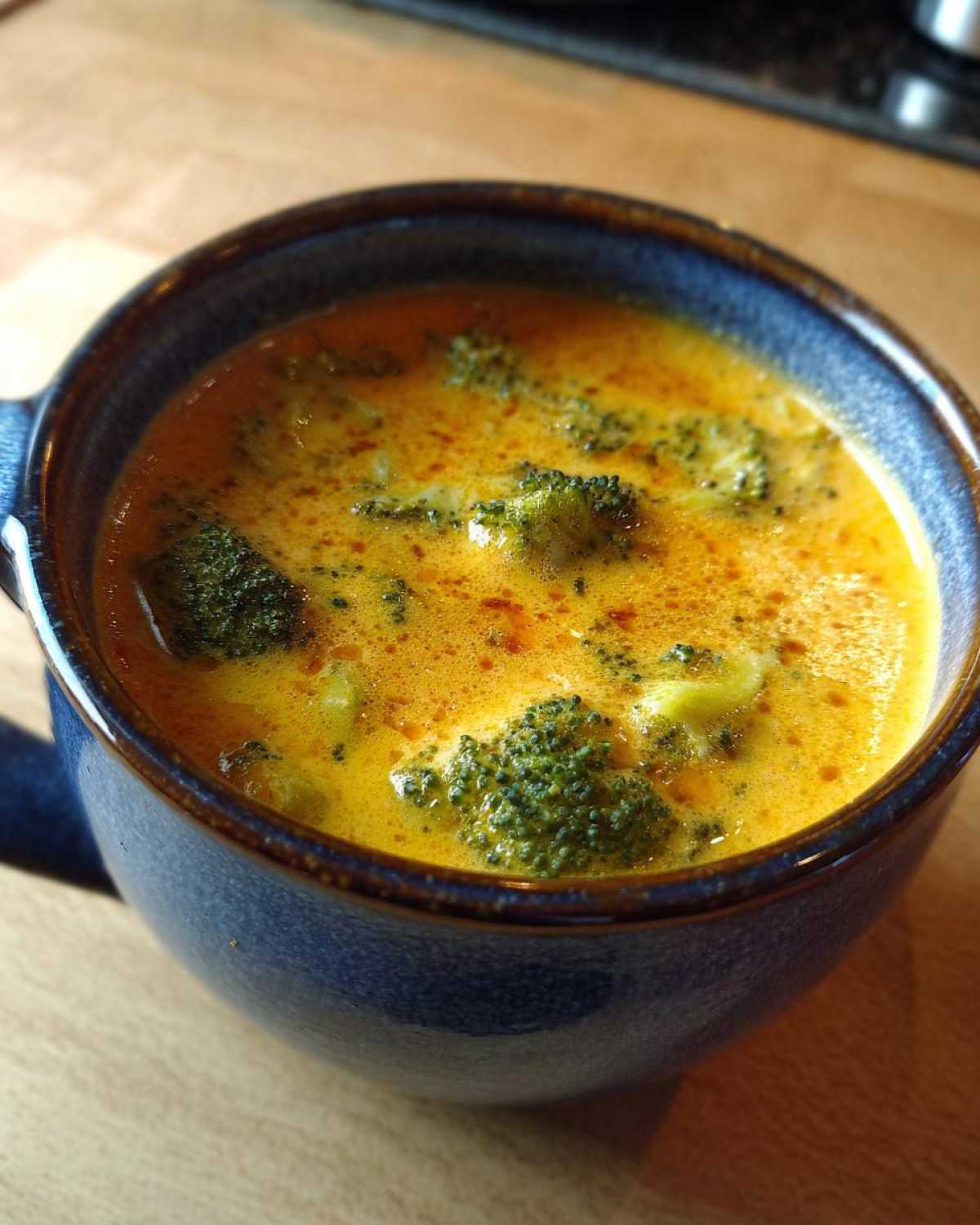 Close-up of creamy, orange-yellow broccoli cheddar soup with visible florets in a rustic blue mug.