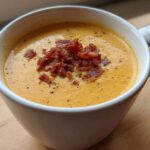 Close-up of creamy, orange-hued cauliflower soup topped generously with crispy bacon bits and black pepper in a white mug.