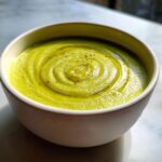 A close-up of vibrant green celery soup, garnished with a swirl of olive oil and cracked black pepper.