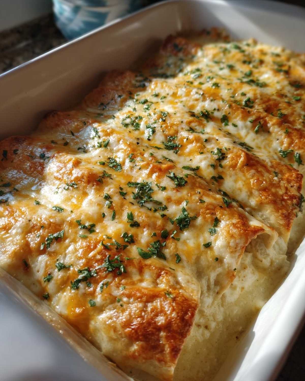 Close-up of creamy chicken enchiladas baked in a white dish, topped with bubbly, melted cheese and parsley.