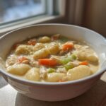 A close-up of a hearty bowl of creamy chicken gnocchi soup with carrots and celery, illuminated by window light.