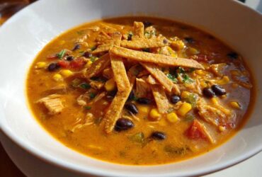 Close-up of a bowl of creamy chicken tortilla soup, topped with crispy tortilla strips, black beans, and corn.