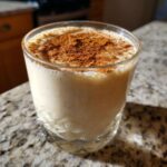 A glass of creamy, pale yellow coquito topped generously with ground cinnamon.