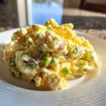 A close-up of a creamy, yellow egg salad recipe portioned onto a white plate, showing chunks of egg, green onions, and red onion.