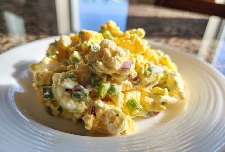 A close-up of a creamy, yellow egg salad recipe portioned onto a white plate, showing chunks of egg, green onions, and red onion.