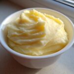 A small white bowl filled with fluffy, yellow creamy mashed potatoes, highlighted by natural sunlight.