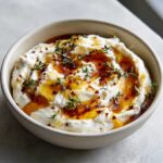 A close-up of creamy ricotta dip topped with golden honey, chili flakes, and fresh thyme sprigs.