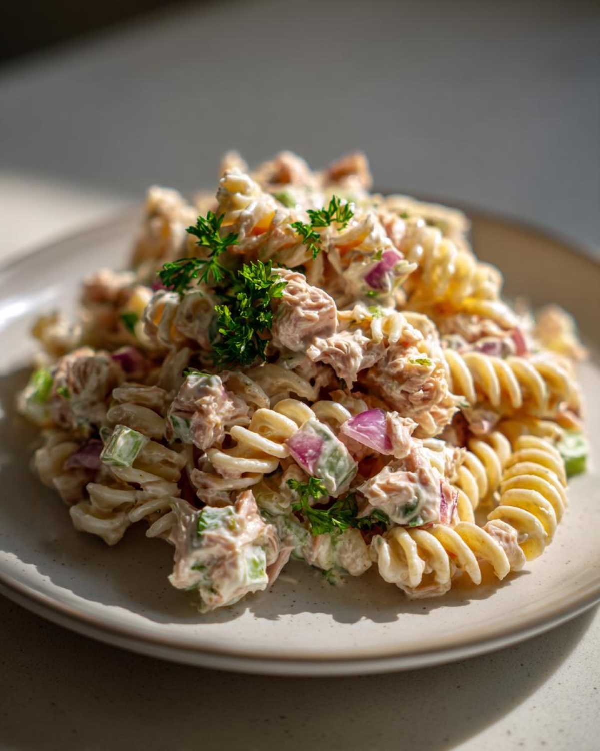 A close-up of creamy tuna pasta salad made with rotini pasta, red onion, and celery, garnished with parsley.