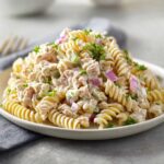 Close-up of a serving of creamy tuna pasta salad made with rotini pasta, chunks of tuna, red onion, and celery, garnished with parsley.