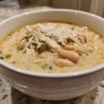 A close-up of a white bowl filled with creamy white chicken chili recipe, topped with shredded cheese.