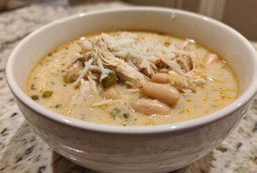 A close-up of a white bowl filled with creamy white chicken chili recipe, topped with shredded cheese.