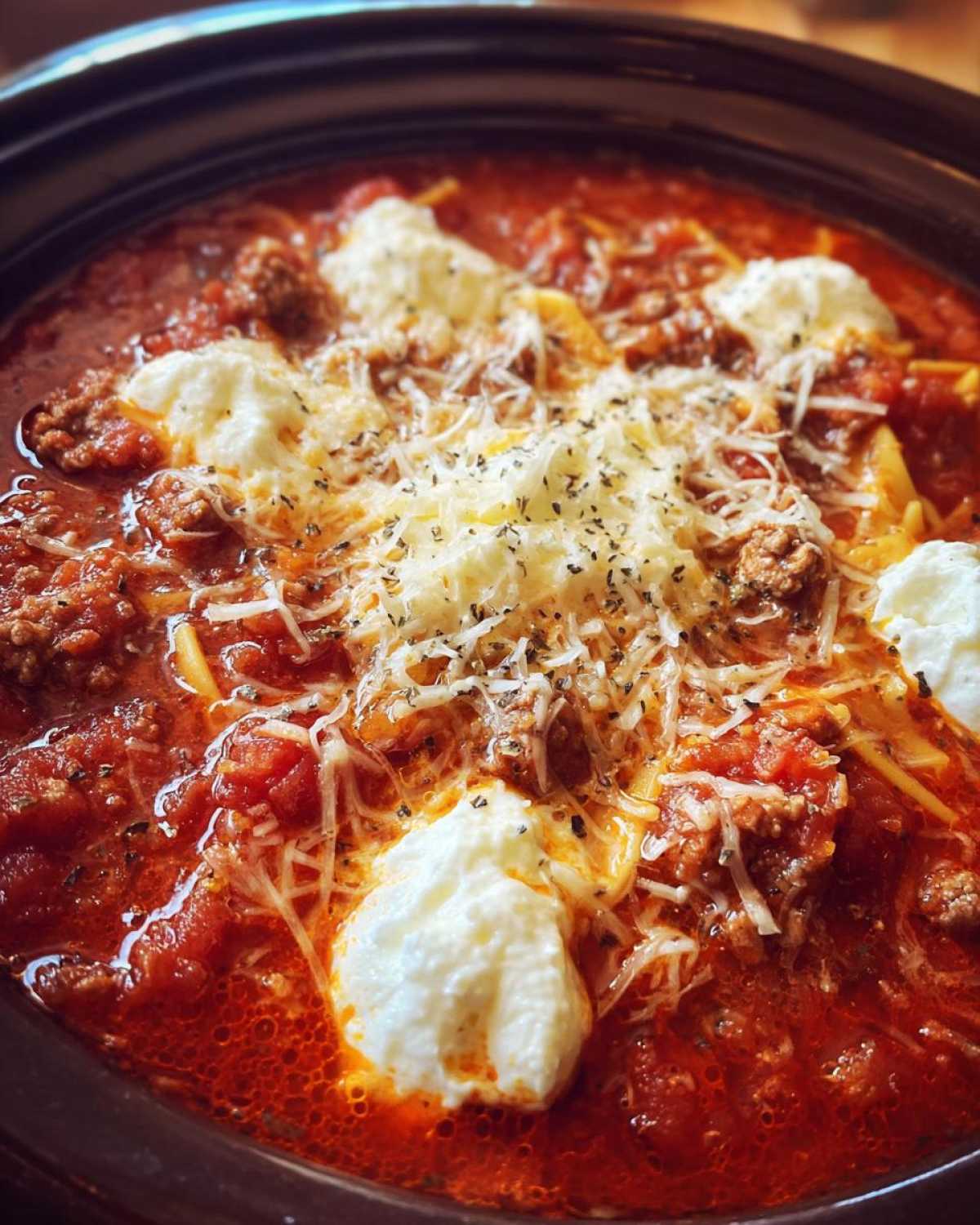 A close-up view of rich, red crockpot lasagna soup topped with dollops of ricotta, shredded mozzarella, and herbs.