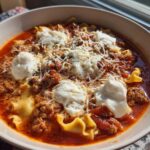 A bowl of rich, red crockpot lasagna soup topped with dollops of ricotta cheese, shredded parmesan, and herbs.
