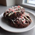 Two rich double chocolate peppermint cookies stacked on a white plate, drizzled with white chocolate and topped with crushed candy canes.