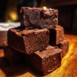 A stack of rich, dark chocolate fudge squares resting on a wooden surface, highlighting the texture of the fudge.