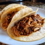 Three soft corn tortillas filled generously with saucy, shredded chicken tacos, served on a white plate.