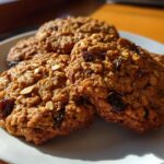 Close-up of several golden brown oatmeal raisin cookies stacked slightly on a white plate, catching warm sunlight.