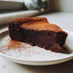 A decadent slice of flourless chocolate torte dusted heavily with cocoa powder, served on a white plate.