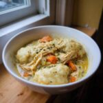 A close-up of a bowl of creamy chicken and dumpling stew, topped with shredded chicken, carrots, and herbs.