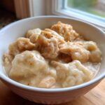 Close-up of a hearty bowl of creamy chicken and dumpling stew, served in a white bowl near a window.