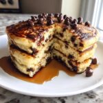 A tall stack of fluffy chocolate chip pancakes cut to show the interior, drizzled with syrup and topped with chocolate chips.