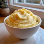 A white bowl overflowing with fluffy, rich creamy mashed potatoes, set near a bright window.