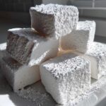 A stack of thick, white homemade marshmallows generously coated in powdered sugar on a white plate.