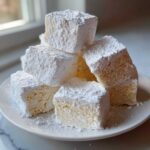 A stack of square, fluffy homemade marshmallows heavily dusted with powdered sugar on a white plate.