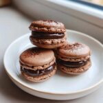 Three decadent chocolate macarons filled with rich ganache, dusted with cocoa powder, sitting on a white plate.